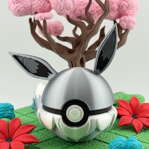 May include: A 3D-printed sculpture of a silver and white ball with black details and rabbit ears, set against a brown tree with pink blossoms and red and blue floral accents on a green grid.