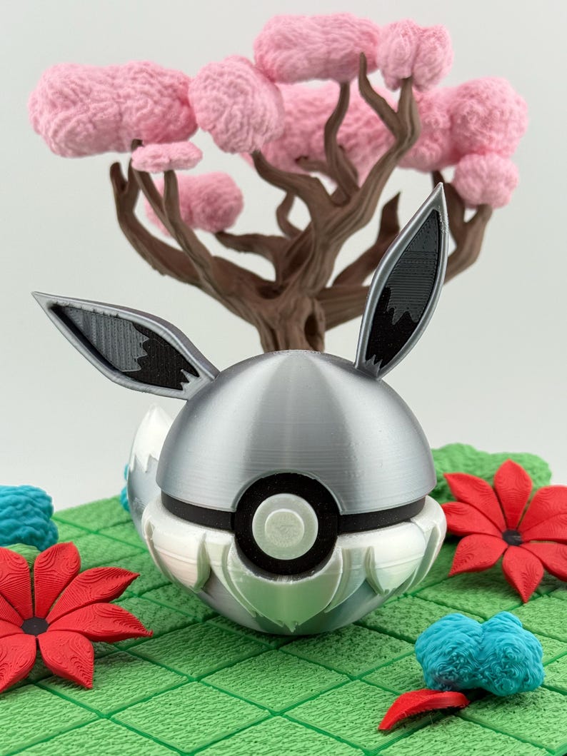 May include: A 3D-printed sculpture of a silver and white ball with black details and rabbit ears, set against a brown tree with pink blossoms and red and blue floral accents on a green grid.