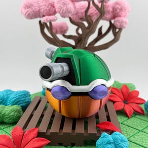 May include: A colorful, 3D-printed toy resembling a character from a video game. The toy features a green shell, orange body, white accents, and purple wheels. A miniature tree with pink blossoms is in the background.