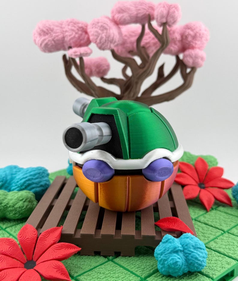 May include: A colorful, 3D-printed toy resembling a character from a video game. The toy features a green shell, orange body, white accents, and purple wheels. A miniature tree with pink blossoms is in the background.