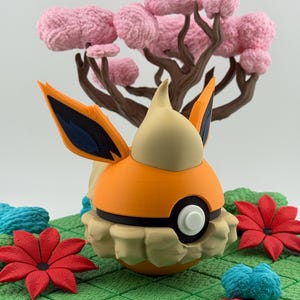 May include: A 3D-printed orange and cream-colored character figure with black accents, resembling a cartoon creature. It is set on a green grid with red and blue floral accents and a pink tree in the background.