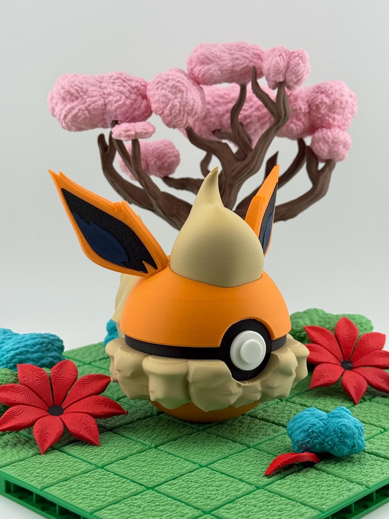 May include: A 3D-printed orange and cream-colored character figure with black accents, resembling a cartoon creature. It is set on a green grid with red and blue floral accents and a pink tree in the background.