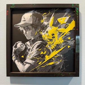 Könnte beinhalten: Gerahmtes Kunstwerk mit einer Graustufen-Illustration einer Person mit Kappe, die einen Pokéball hält, mit einem leuchtend gelben Pikachu, das aus der Seite platzt. Das Kunstwerk ist vor einem schwarzen Hintergrund platziert und in einem dunkelbraunen Rahmen gerahmt.