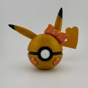 May include: A golden-yellow, spherical decorative item with black and white accents, orange details, and a pink bow. The item has two pointed ears with black tips and a tail-like shape. The design is reminiscent of a popular animated character.
