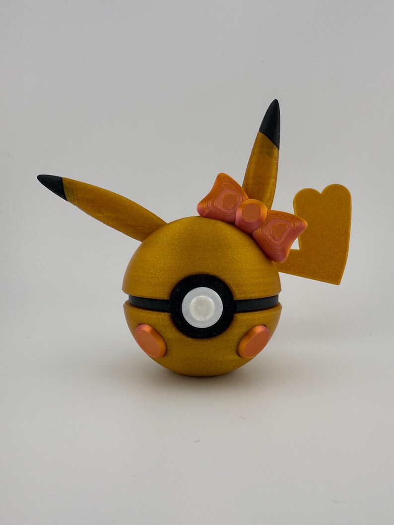 May include: A golden-yellow, spherical decorative item with black and white accents, orange details, and a pink bow. The item has two pointed ears with black tips and a tail-like shape. The design is reminiscent of a popular animated character.