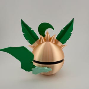 May include: A 3D-printed, spherical object with a metallic gold finish, featuring green leaf-like appendages and a spiky crown. A black band encircles the sphere. The design is reminiscent of a stylized plant or fruit.