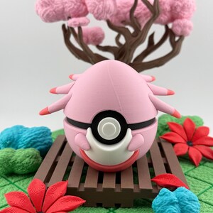 May include: A pink, spherical figurine with black and white accents, resembling a character from a popular animated series. It rests on a brown wooden platform, surrounded by red flowers, blue and green decorative elements, and a pink tree.