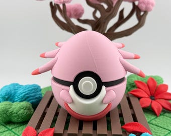 Op de afbeelding: Een roze, bolvormig beeldje met zwarte en witte accenten, dat lijkt op een personage uit een populaire animatieserie. Het staat op een bruin houten platform, omringd door rode bloemen, blauwe en groene decoratieve elementen en een roze boom.