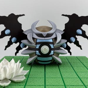 May include: A detailed, futuristic-looking sculpture with a central orb-like body featuring blue and gray accents, flanked by black wing-like structures with blue details. A white lotus flower sits in front of the sculpture on a green surface.