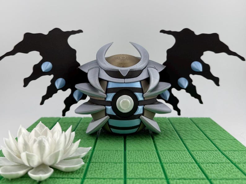 May include: A detailed, futuristic-looking sculpture with a central orb-like body featuring blue and gray accents, flanked by black wing-like structures with blue details. A white lotus flower sits in front of the sculpture on a green surface.