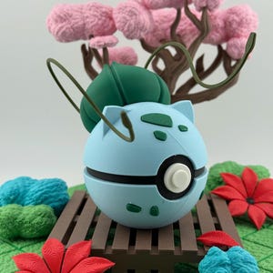 May include: A light blue, spherical figure with green details and a white centre, reminiscent of a popular character, rests on a brown wooden platform. The scene includes pink tree-like structures, red flowers, and green foliage.