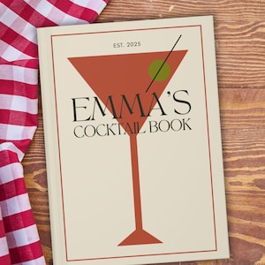 May include: A cream-coloured cocktail book with a red martini glass design on the cover. The book is titled "EMMA'S COCKTAIL BOOK" with "EST. 2025" above the glass. A red and white checked cloth is visible on the left.
