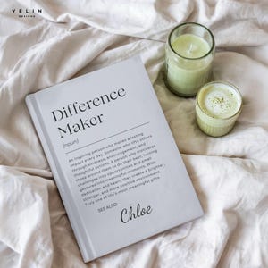 Może przedstawiać: Biała książka zatytułowana "Difference Maker" z definicją i imieniem "Chloe" jest wyświetlana na białym prześcieradle. Obok książki znajdują się dwa szklane pojemniki, jeden z zieloną świecą, a drugi z zielonym napojem.
