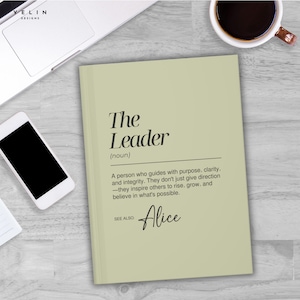 May include: A sage green notebook with the text "The Leader" and a definition. The notebook also includes the name "Alice" and is next to a smartphone and a cup of coffee.