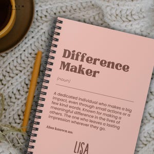 May include: A spiral-bound notebook with a light pink cover. The cover features the words "Difference Maker" in a bold, brown font, with a definition below. A pen rests beside the notebook. The notebook is on a white knitted surface.