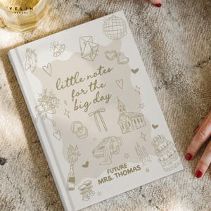 May include: A white hardback notebook featuring gold line art illustrations of wedding icons including a church, cake, champagne, and rings. The text reads "little notes for the big day" and "FUTURE MRS. THOMAS" in a gold script and block font.