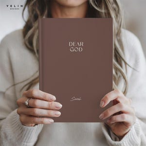 May include: A brown hardcover journal featuring the text DEAR GOD in a serif font at the top and the name Sarah in a cursive script font at the bottom. The cover has a smooth, matte finish and the book is held upright in a vertical orientation.