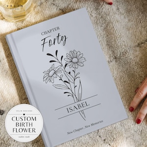 Peut inclure: Un livre à couverture rigide gris clair avec une illustration au trait noir représentant un bouquet de fleurs. Le texte indique CHAPTER Forty, ISABEL et New Chapter, New Memories. Un logo circulaire affiche VELIN DESIGNS CUSTOM BIRTH FLOWER.