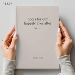 May include: A beige notebook with the text "notes for our happily ever after" and the names "Chloe & James" printed on the cover. The notebook is held by two hands with manicured nails.