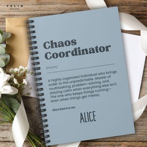 以下が含まれることがあります： 「Chaos Coordinator」の文字が大きくダークグレーで書かれた、水色のスパイラル綴じノート。用語の定義が下に印刷され、「ALICE」という名前も記載されています。ノートは木製の表面に置かれています。