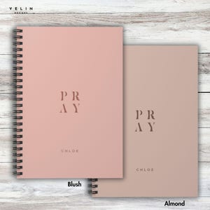 Könnte beinhalten: Gezeigt werden zwei spiralgebundene Notizbücher, eines in Blush-Rosa und das andere in Mandel. Das Wort "PRAY" ist vertikal in einer minimalistischen Schriftart auf jeder Hülle aufgedruckt, darunter der Name "CHLOE". Die Notizbücher liegen auf einer hellen Holzmaserung.