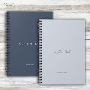 Puede incluir: Dos cuadernos con espiral, uno azul marino y otro gris claro, se muestran sobre una superficie de madera. El cuaderno azul marino tiene las palabras "CUSTOM TEXT" impresas en la portada. El cuaderno gris claro también tiene las palabras "CUSTOM TEXT" impresas en la portada.