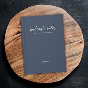 May include: A slate blue notebook with the words "podcast notes" in a cursive font. Below the title are the words "IDEAS | PLANNING | RECORDING". The name "MATTHEW" is printed at the bottom. The notebook rests on a wooden serving board.