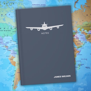 May include: A dark grey notebook featuring a white aeroplane illustration and the word "NOTES". The name "JAMES WILSON" is printed at the bottom. The notebook is set against a world map background.