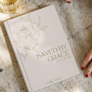 May include: A light beige prayer journal with a dark brown line art illustration of flowers on the cover. The text reads SAVED BY GRACE prayer journal in a clean sans-serif font, with the name Ashley Wilson printed in a cursive script at the bottom.