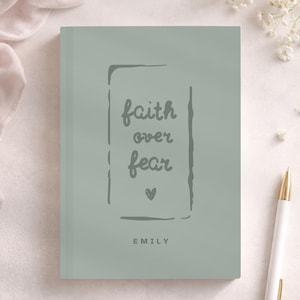 Puede incluir: Un diario de tapa dura color verde salvia que presenta un borde rectangular gris oscuro con el texto faith over fear escrito en una fuente cursiva en su interior, seguido de un pequeño icono de corazón y el nombre EMILY en mayúsculas.