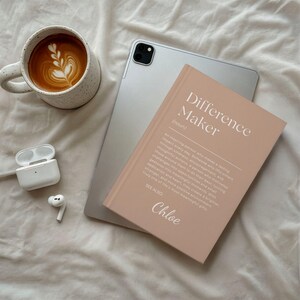 Może przedstawiać: Jasnor&oacute;żowy notatnik z napisem "Difference Maker" i imieniem "Chloe" obok srebrnego tabletu, filiżanki kawy z latte art i pary białych bezprzewodowych słuchawek.