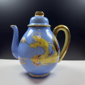 May include: A light blue porcelain teapot featuring a gold-painted dragon motif and a depiction of a mountain. The teapot has a curved spout, a gold-colored handle, and a matching lid with a gold finial. Gold trim accents the base and rim.