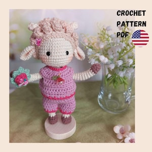 May include: A crocheted sheep doll wearing a pink outfit and holding a flower. The doll has a light pink head and ears, with a small pink flower on its head. The text "CROCHET PATTERN PDF" is visible in the upper right corner.