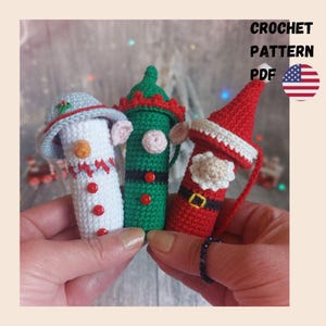 Könnte beinhalten: Drei handgefertigte gehäkelte Weihnachtsschmuckstücke: ein Schneemann, ein Elf und der Weihnachtsmann. Der Schneemann ist weiß mit blauem Hut, der Elf ist grün und der Weihnachtsmann ist rot. Die Ornamente werden in den Händen einer Person gehalten. Der Text auf dem Bild lautet "CROCHET PATTERN PDF".