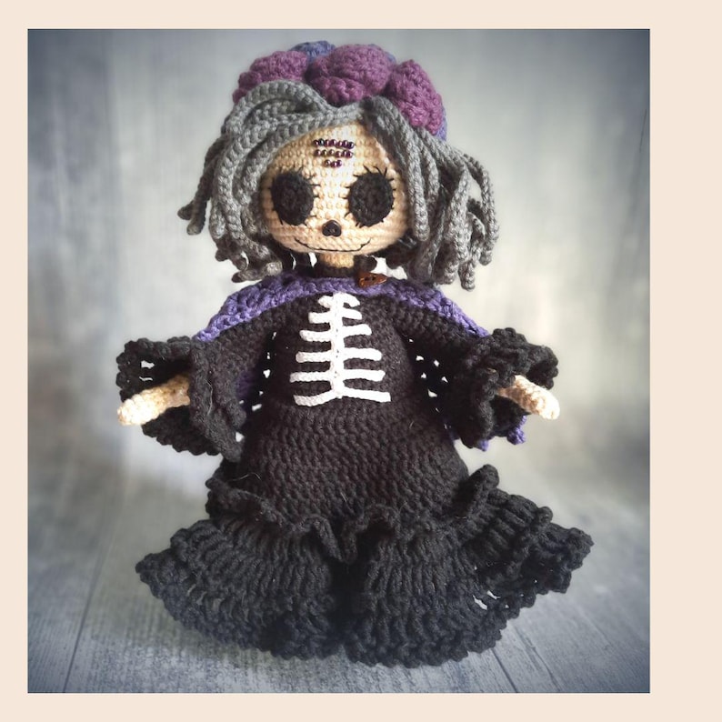 May include: A handmade crochet doll with a spooky theme. The doll has gray hair, a purple and gray hat, and a black dress with a white skeleton design. The doll's face has black eyes and stitched details.