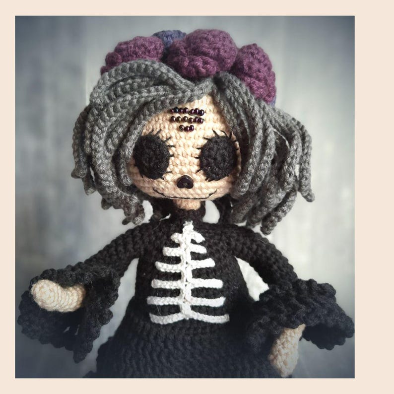 May include: A handmade crochet doll with a skeleton design. The doll has gray hair, a purple flower crown, and a black dress with a white skeleton design. The doll's face is beige with black eyes and a small nose.
