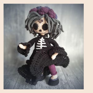 May include: Crocheted doll with a skeleton design on a black dress. The doll has gray hair, a purple headpiece, and purple and black shoes. The doll's arms and hands are beige. The doll is standing on a gray wooden surface.