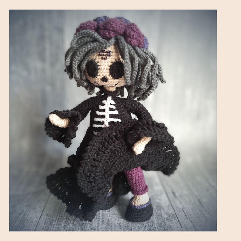 May include: Crocheted doll with a skeleton design on a black dress. The doll has gray hair, a purple headpiece, and purple and black shoes. The doll's arms and hands are beige. The doll is standing on a gray wooden surface.