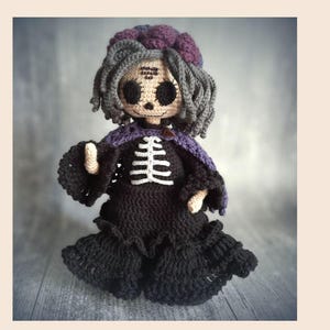 May include: A handmade crochet doll with a skeleton design. The doll has gray hair, a purple shawl, and a black dress. The skeleton is white, and the doll has large, dark eyes. The doll is a unique, handcrafted item.