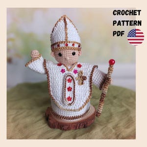 May include: A crocheted figure of a religious leader, wearing a white robe with gold trim and a matching hat. The figure holds a staff with a red orb. The text "CROCHET PATTERN PDF" is visible in the image.