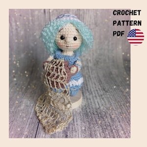 Puede incluir: Una muñeca de ganchillo hecha a mano con cabello azul y un vestido azul, sosteniendo un objeto marrón y rosa, y un encaje dorado. La imagen incluye el texto "CROCHET PATTERN PDF" y la bandera estadounidense.