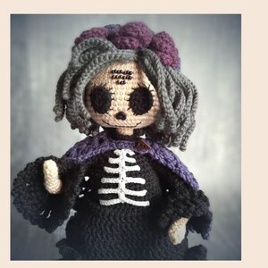 May include: A handmade crochet doll with a skeleton design. The doll has gray hair, a purple flower, and a black dress with a white rib cage. The doll's face is beige with black eyes and a stitched smile. A purple shawl is around the doll's shoulders.
