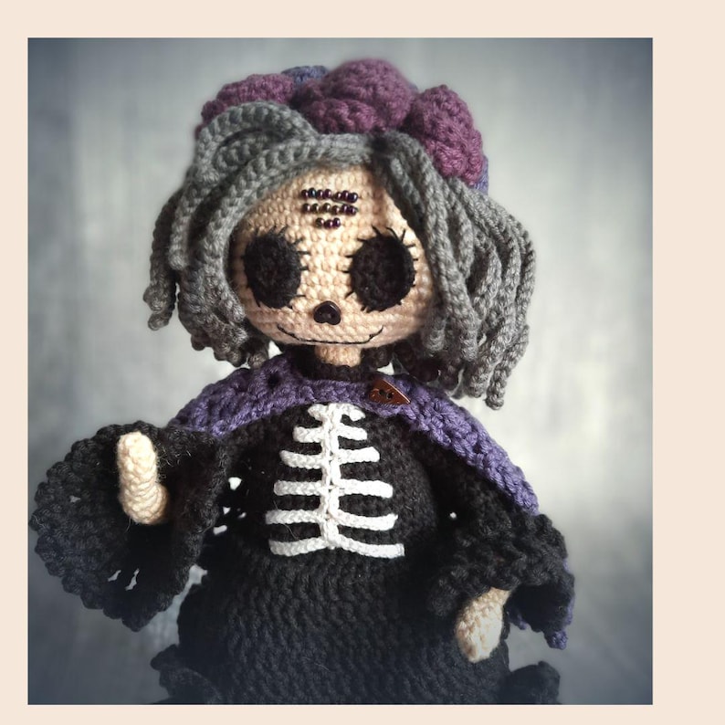 May include: A handmade crochet doll with a skeleton design. The doll has gray hair, a purple flower, and a black dress with a white rib cage. The doll's face is beige with black eyes and a stitched smile. A purple shawl is around the doll's shoulders.