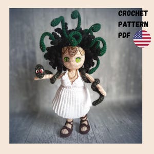 Könnte beinhalten: Gehäkelte Medusa-Puppe mit grünen Augen, schwarzen Haaren und grünen Schlangenhaaren. Sie trägt ein weißes Kleid und braune Sandalen und hält eine kleine Schlange. Das Bild enthält den Text "CROCHET PATTERN PDF" und eine amerikanische Flagge.