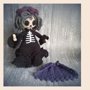 May include: A handmade crochet doll with a black dress and a white skeleton design on the front. The doll has gray hair, a purple floral headpiece, and a matching purple cape. The doll is a unique, handcrafted item.