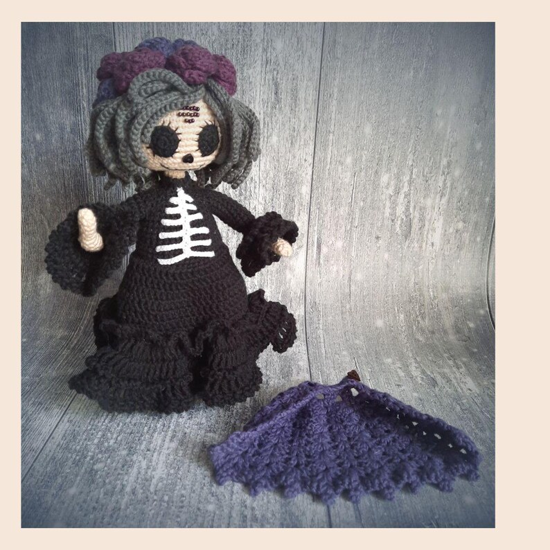 May include: A handmade crochet doll with a black dress and a white skeleton design on the front. The doll has gray hair, a purple floral headpiece, and a matching purple cape. The doll is a unique, handcrafted item.