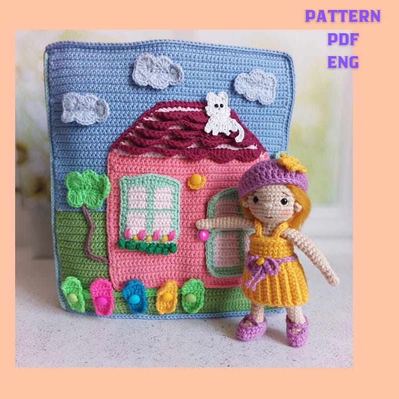 May include: A crocheted house-shaped pillow with a pink house, blue sky, and white clouds. A crocheted doll in a yellow dress and purple hat stands in front of the pillow. The pillow has the text "PATTERN PDF ENG".