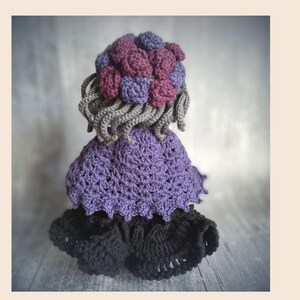 May include: A handmade crochet doll with a purple dress, black skirt, and gray hair. The doll's hair is adorned with purple and pink crochet flowers. The doll is a unique, handcrafted item.