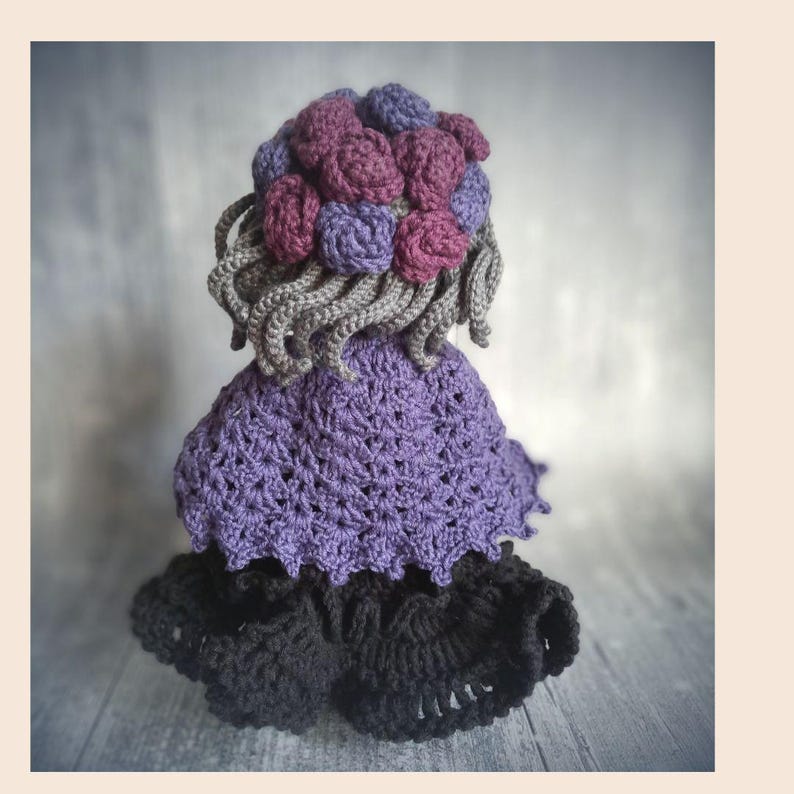 May include: A handmade crochet doll with a purple dress, black skirt, and gray hair. The doll's hair is adorned with purple and pink crochet flowers. The doll is a unique, handcrafted item.