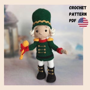 Puede incluir: Muñeco cascanueces de ganchillo hecho a mano con uniforme verde con detalles dorados, botas negras y sombrero a juego. El muñeco sostiene un micrófono amarillo y rojo. La imagen incluye el texto "CROCHET PATTERN PDF" y una bandera americana.